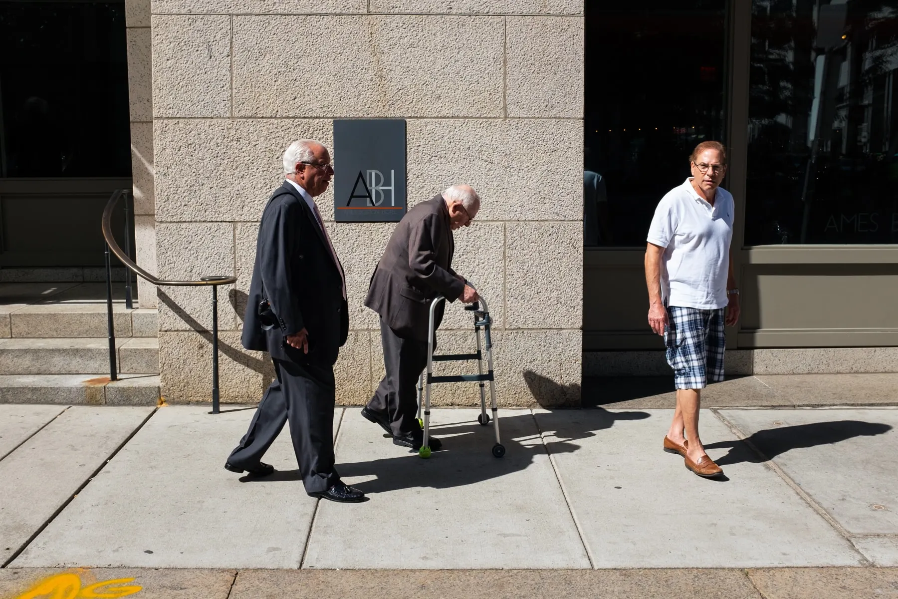 Two older men, one using a walker, pass a man watching from a doorway