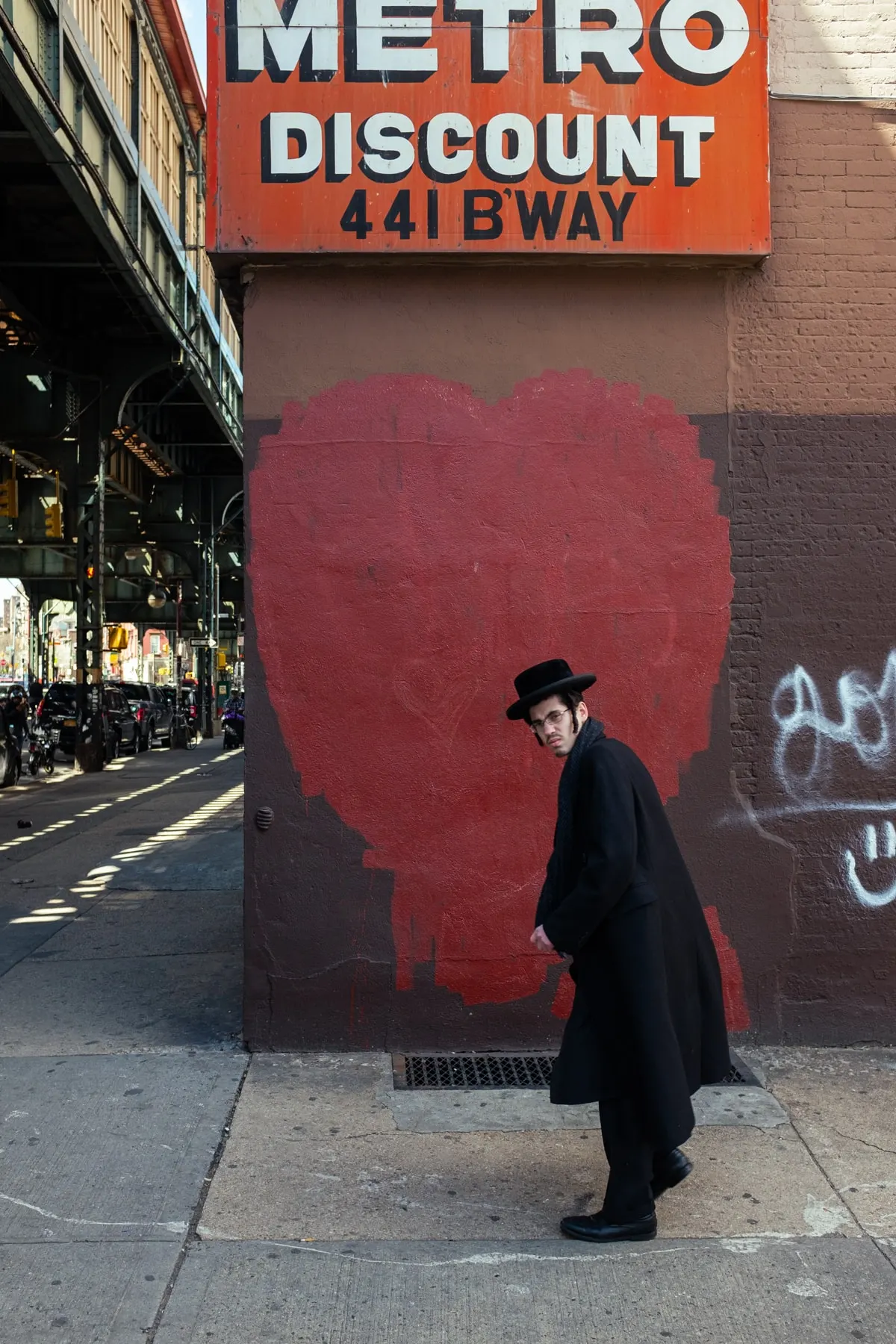 Man in a black coat and hat glances back in front of a red heart painted on brick