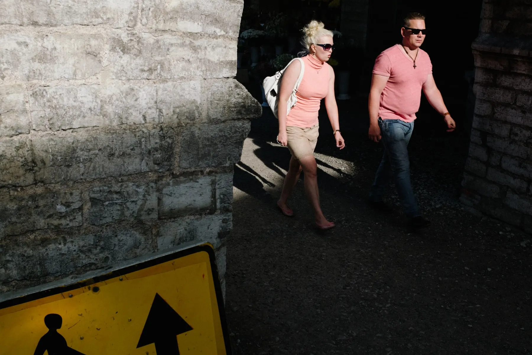 Couple dressed in pink walk out of shadow beside a rough stone wall