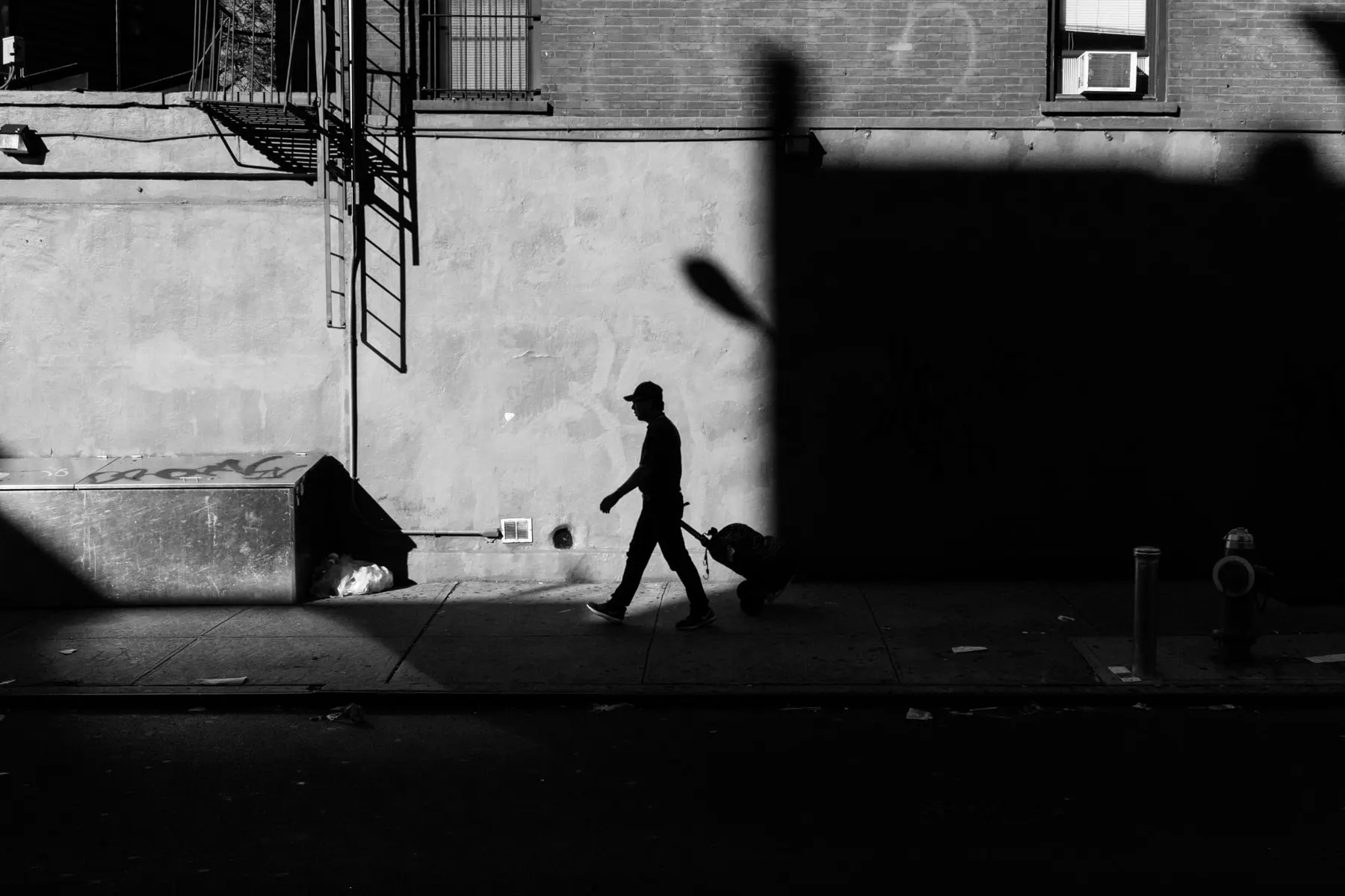 Man wheels a suitcase along a sunlit strip of sidewalk beside deep shadow