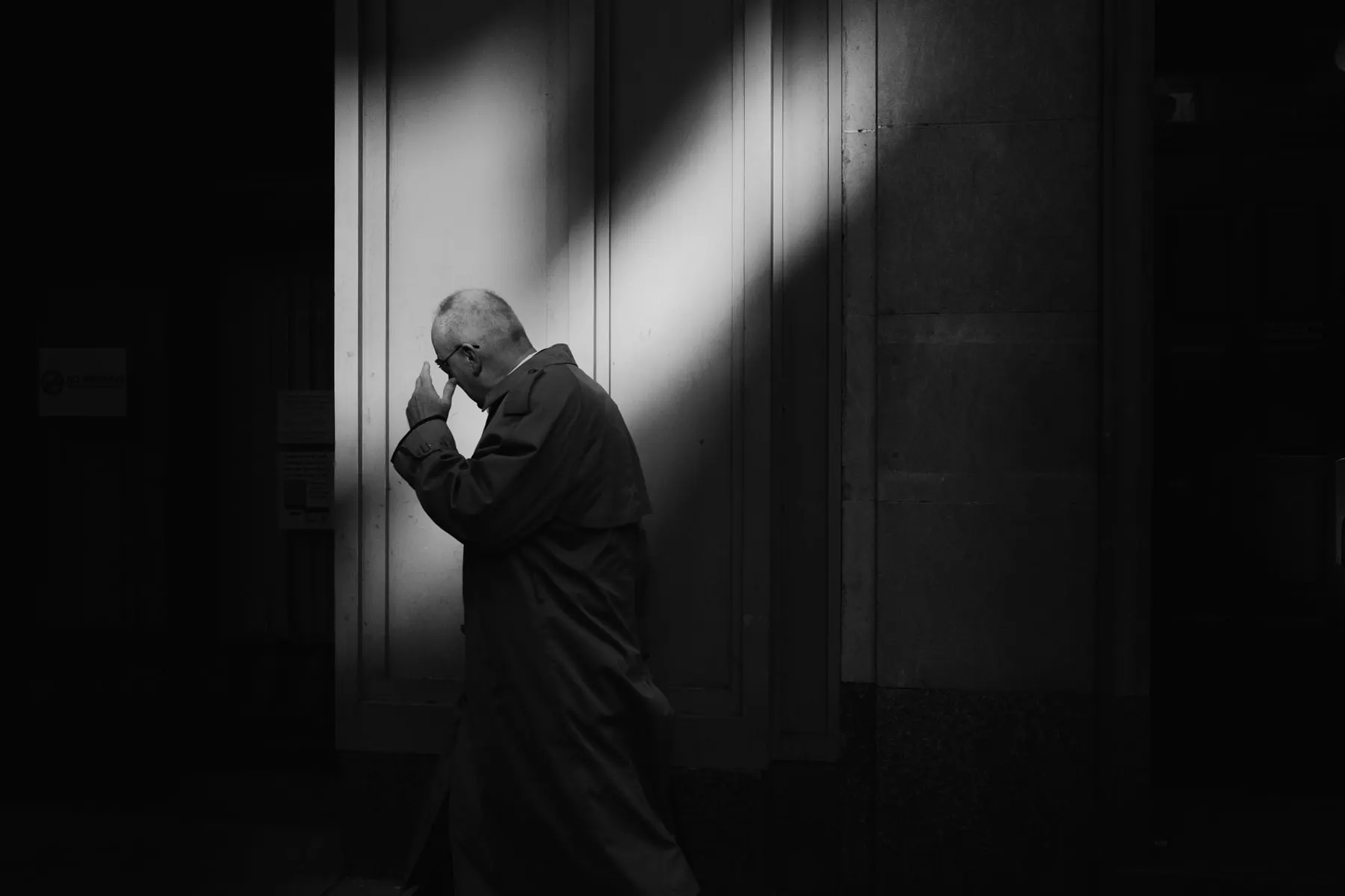 Older man in a coat covers his face in a shaft of light against a dark wall