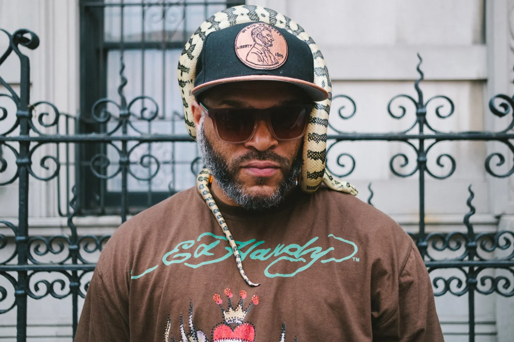 Man in sunglasses and a snake-print cap in front of an iron fence